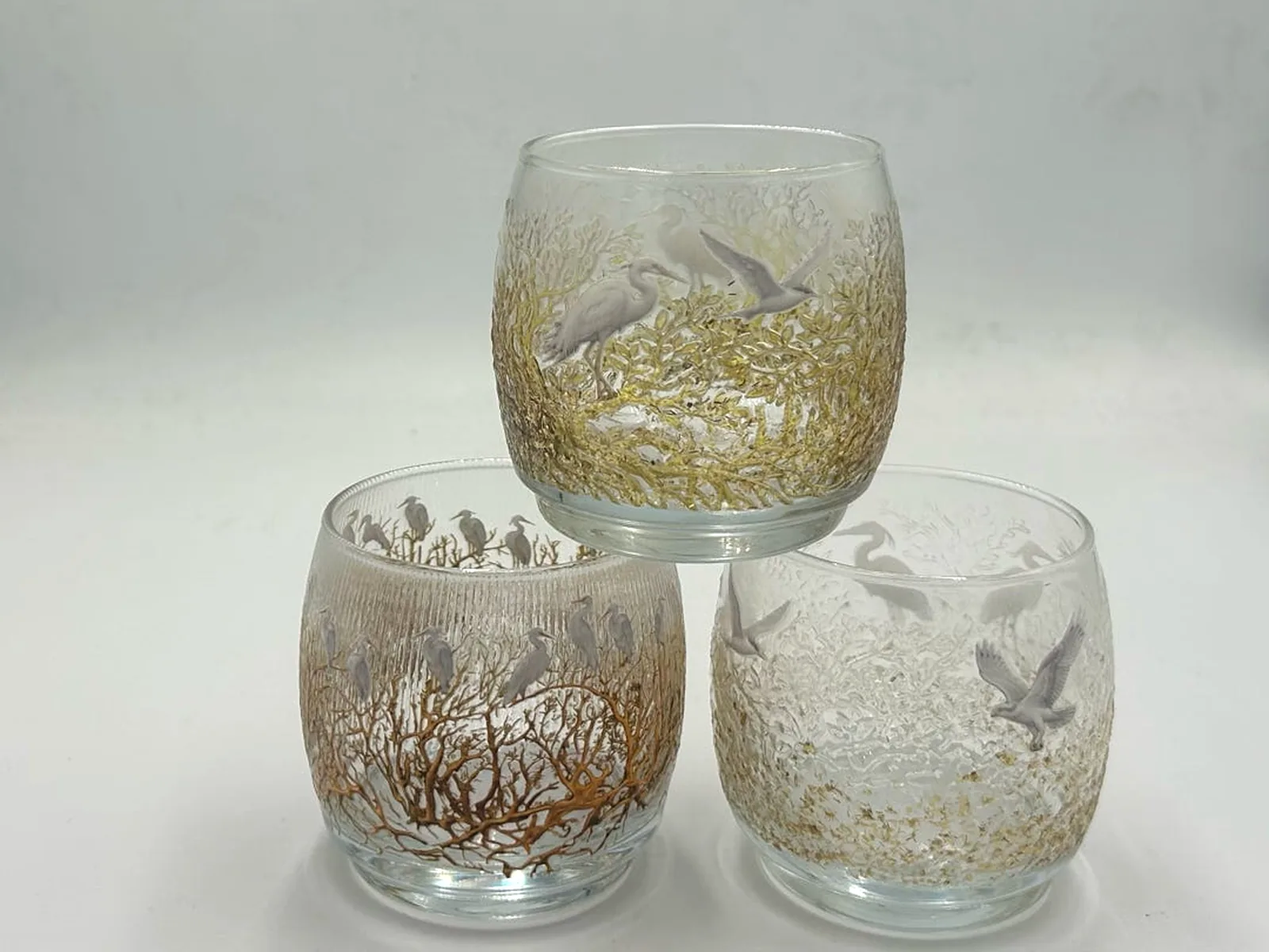 Three clear glass roly-poly tumblers stacked in a pyramid, each printed all the way round with white egrets among gold and copper coral-like branches