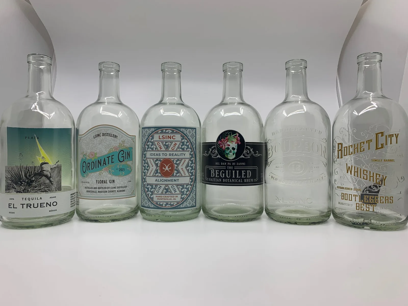 Six clear glass spirit bottles in a row, each printed directly with a different label-style design: a full-colour photo panel, pastel and teal panels, a black panel with a skull and flowers, a design printed in clear for an embossed look, and gold-coloured lettering
