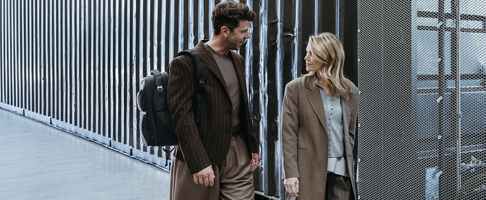 A man and a woman talking as they walk past a slatted metal wall, the man carrying a black DICOTA laptop backpack on one shoulder.