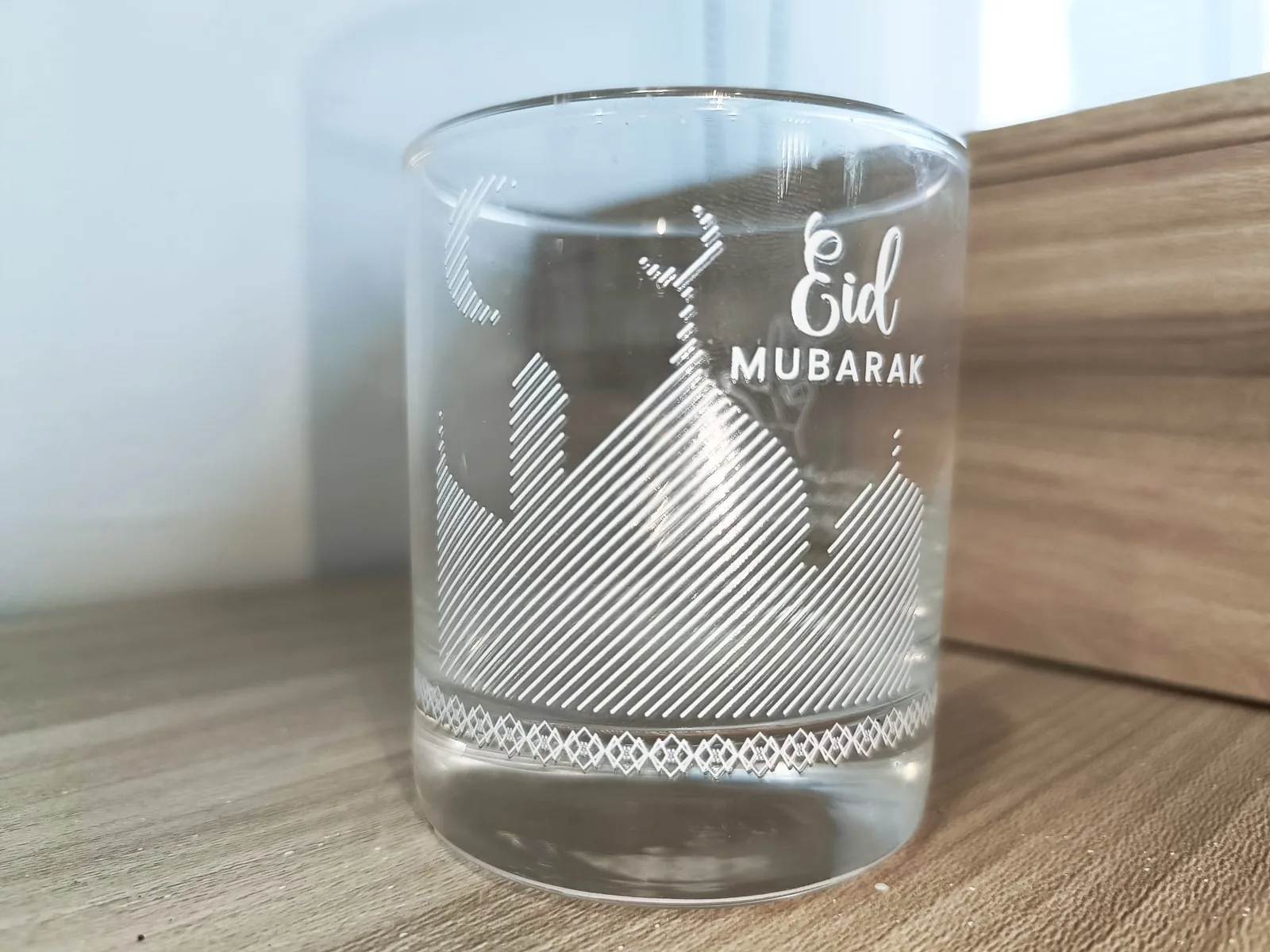 A clear glass rocks tumbler printed in white ink with a mosque silhouette drawn in diagonal lines, a crescent moon, the words Eid Mubarak and a geometric border near the base