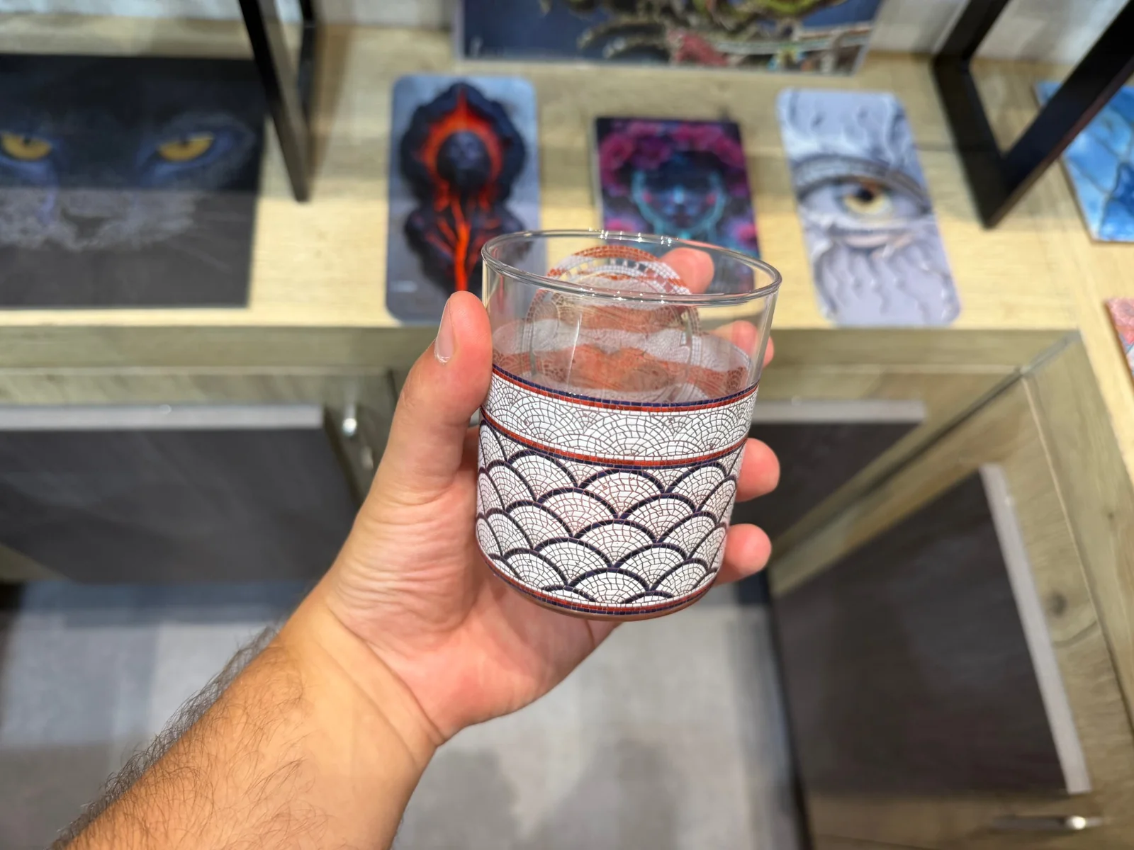 يد تمسك كأساً زجاجياً شفافاً مطبوعاً حول جزئه السفلي بفسيفساء بيضاء من أقواس على شكل حراشف السمك محددة بالكحلي، بين شريطين أحمر وكحلي، مع ترك الجزء العلوي من الزجاج شفافاً