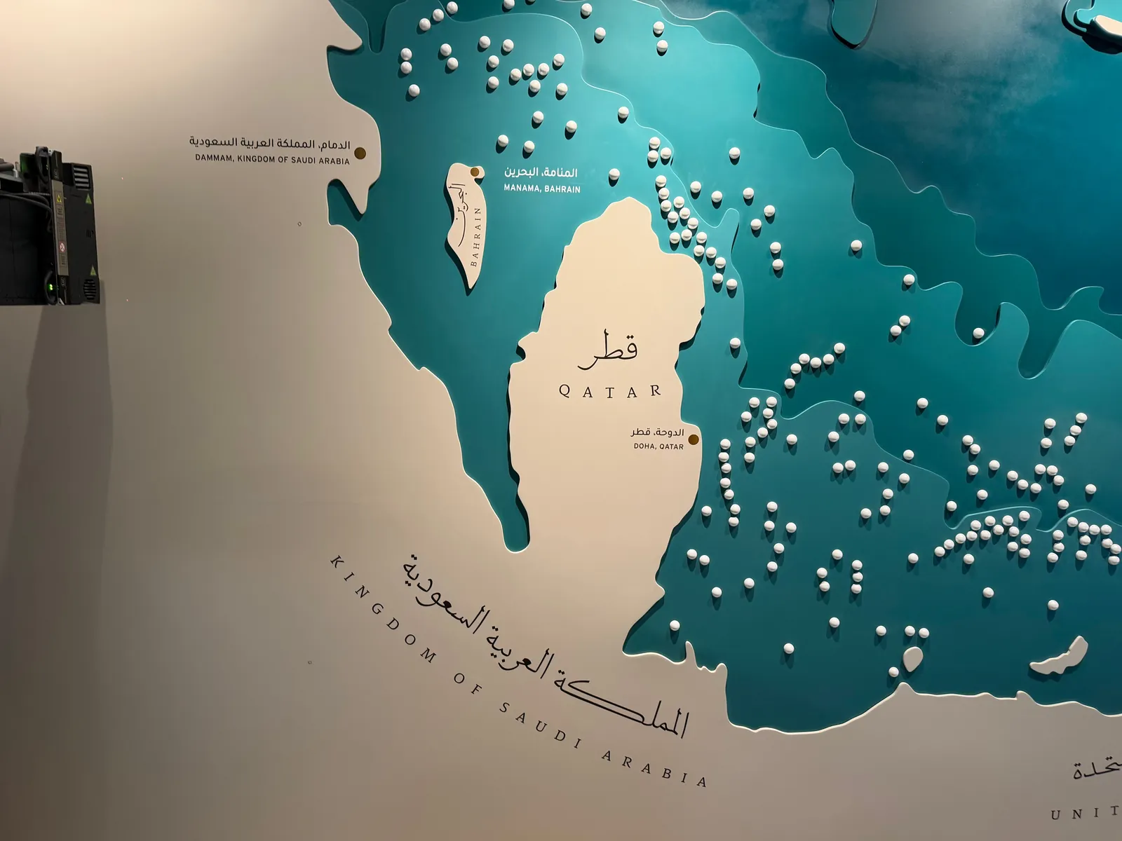 Close-up of an installed museum map panel: raised teal layers, pearl-like spheres standing out from the surface, an island shape printed with the name Bahrain, and the label Manama, Bahrain printed in white