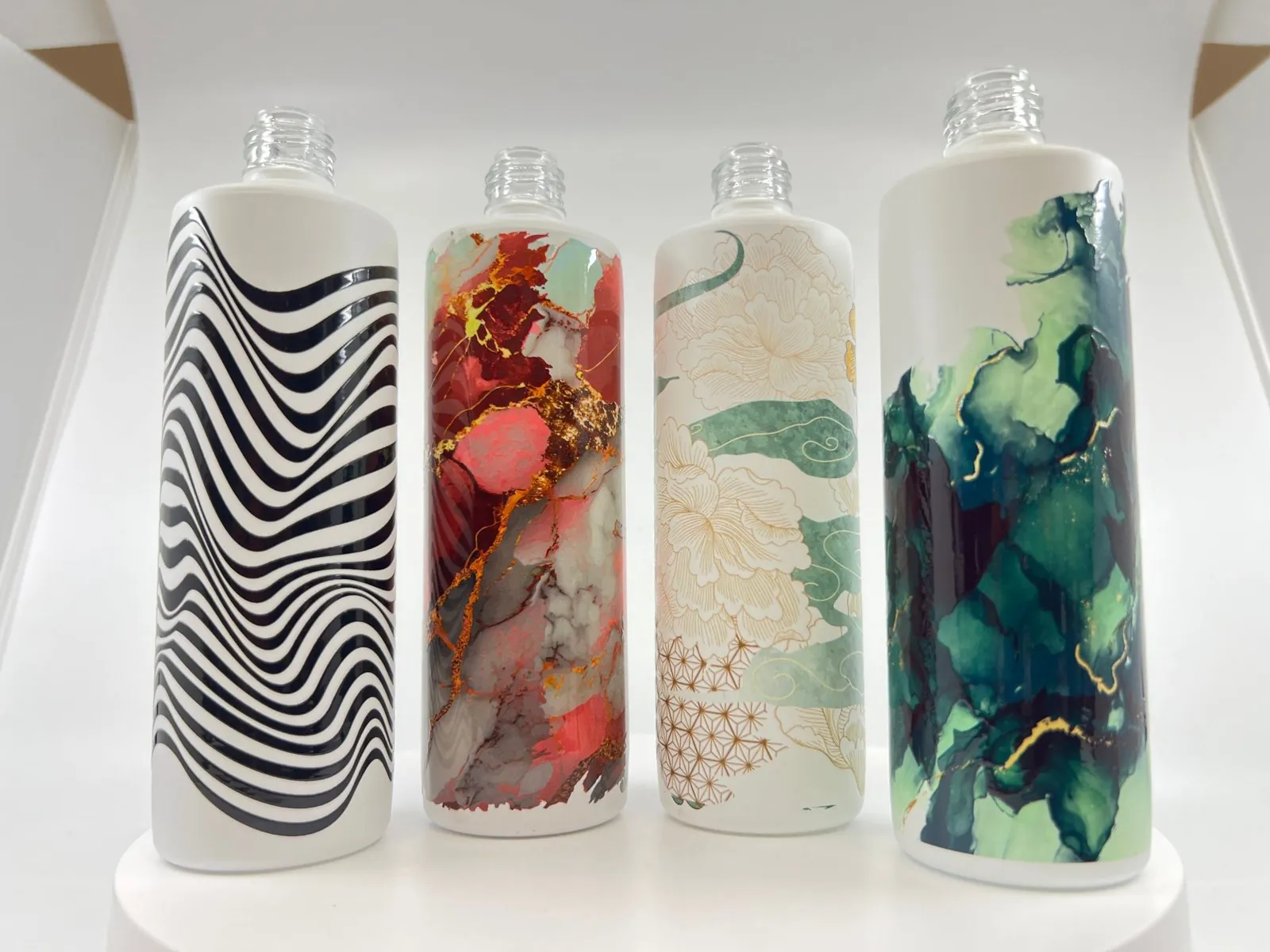 Four white cylindrical bottles printed 360 degrees with different designs: black zebra stripes, red marble, a floral illustration and a green marble pattern