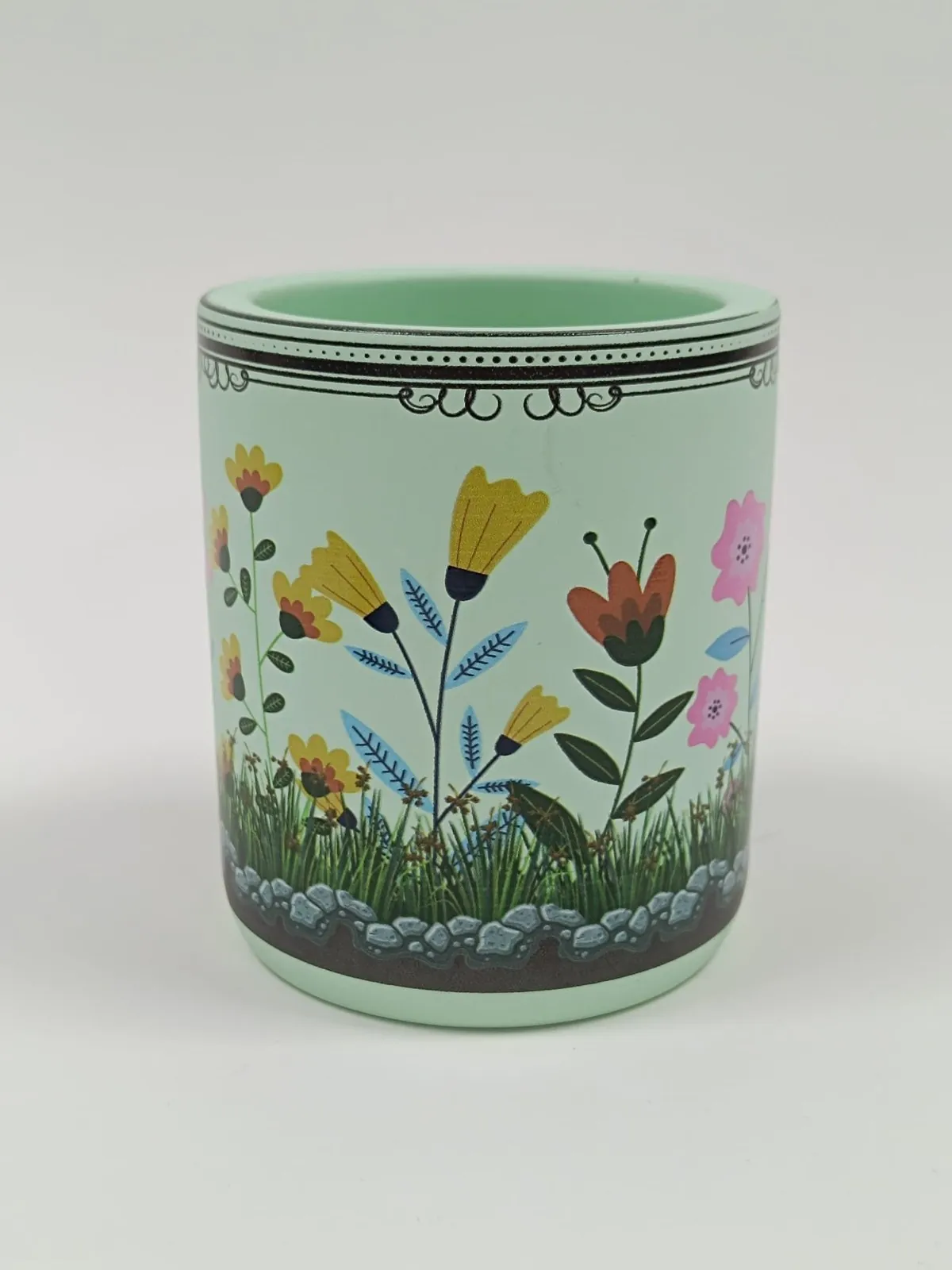 A mint-green candle vessel printed with a full-colour wrap of yellow, orange and pink flowers, grass and stones