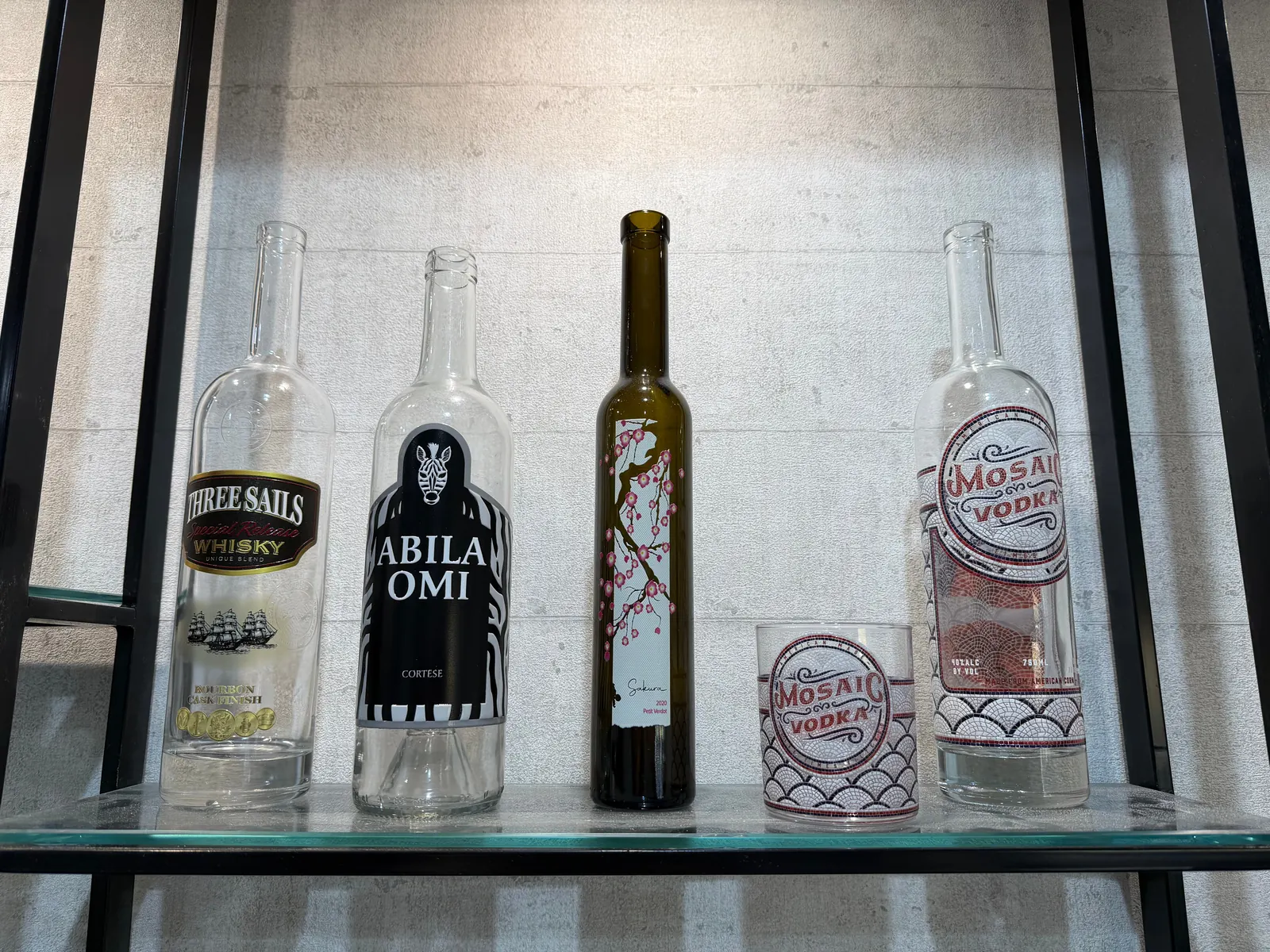 Glass bottles printed directly on an LSINC press, on a sample shelf in Dubai: a whisky-style bottle with a metallic label design and ship illustration, a bottle with a black-and-white zebra design, a dark bottle with cherry blossoms, and a mosaic-pattern bottle with a matching tumbler