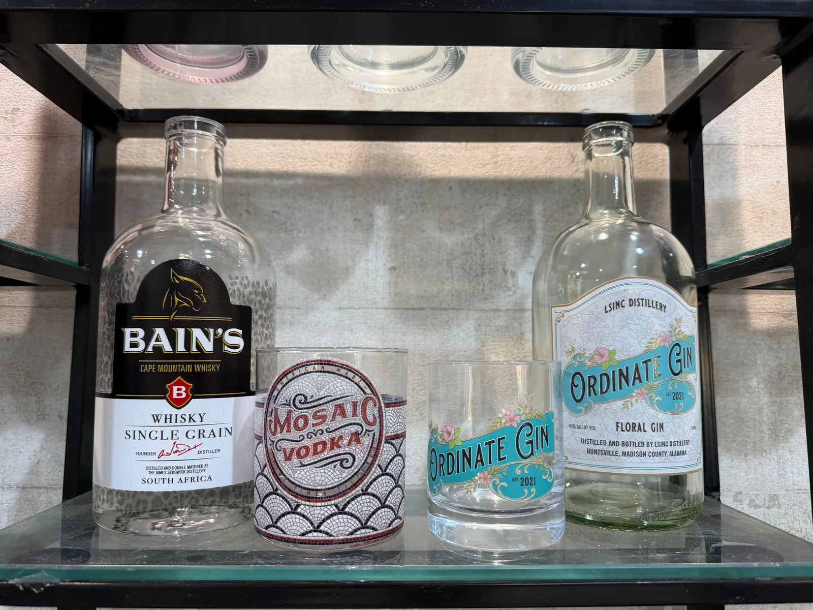 A printed whisky-style glass bottle with a hammered texture and label-style artwork, a tumbler with a mosaic full wrap, a tumbler with a floral label-style design and a matching gin-style bottle, on a sample shelf in Dubai
