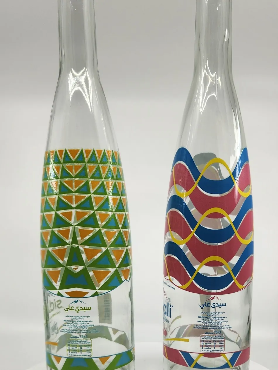 Two clear glass water bottles printed directly with geometric patterns, one in green and orange triangles and one in red, blue and gold waves, with Arabic text at the base
