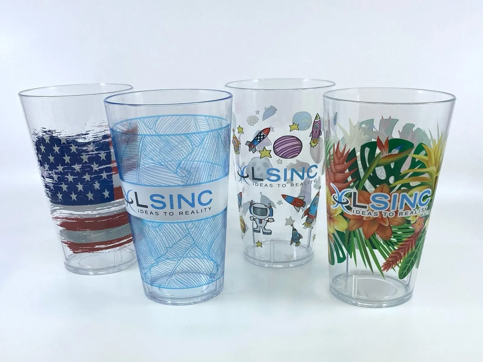 Four clear pint tumblers printed directly by LSINC: a distressed flag, fine blue line work with a frosted band, cartoon rockets and planets, and tropical flowers, three of them with the LSINC logo