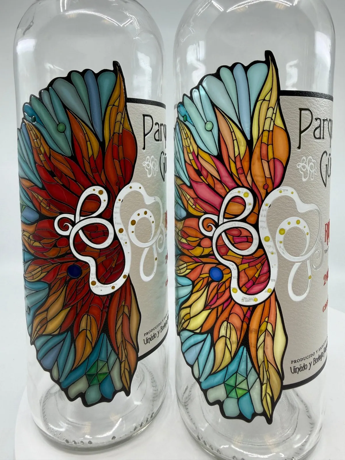 Two clear wine bottles printed directly with a stained-glass style artwork in orange, turquoise and blue around a white label-style panel