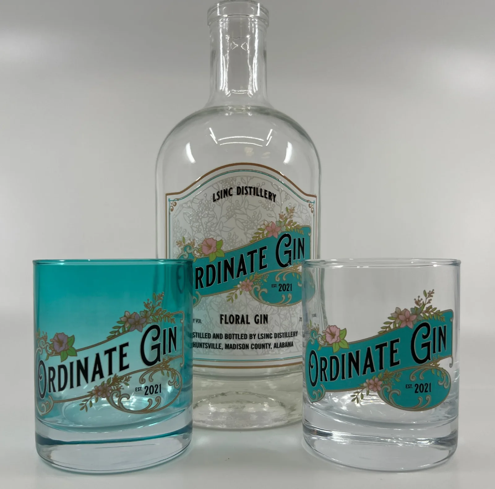 A clear glass gin bottle and two tumblers, one clear and one teal glass, all printed with the same full-colour label design in teal, pink and gold