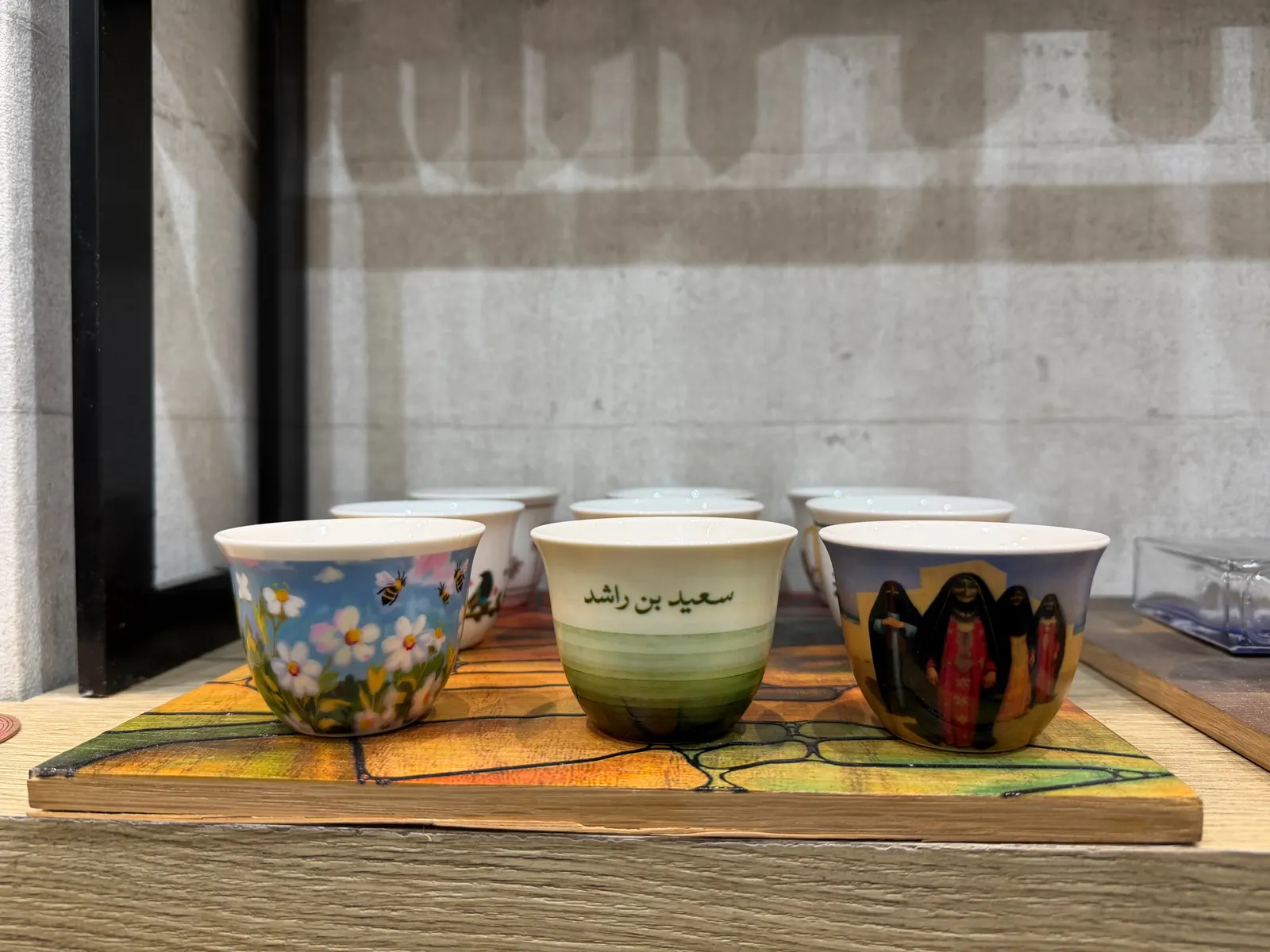 Personalised UV-printed qahwa cups on a wooden display: a cup with bees and daisies, a cup with an Arabic name printed over green stripes, and a cup with a painting of Emirati women in traditional dress