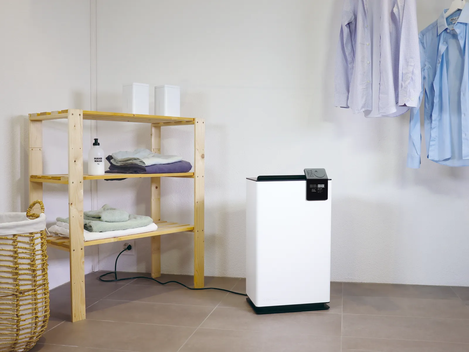 Stadler Form Albert dehumidifier in a laundry room beside a wooden shelf unit and shirts hanging on a rail