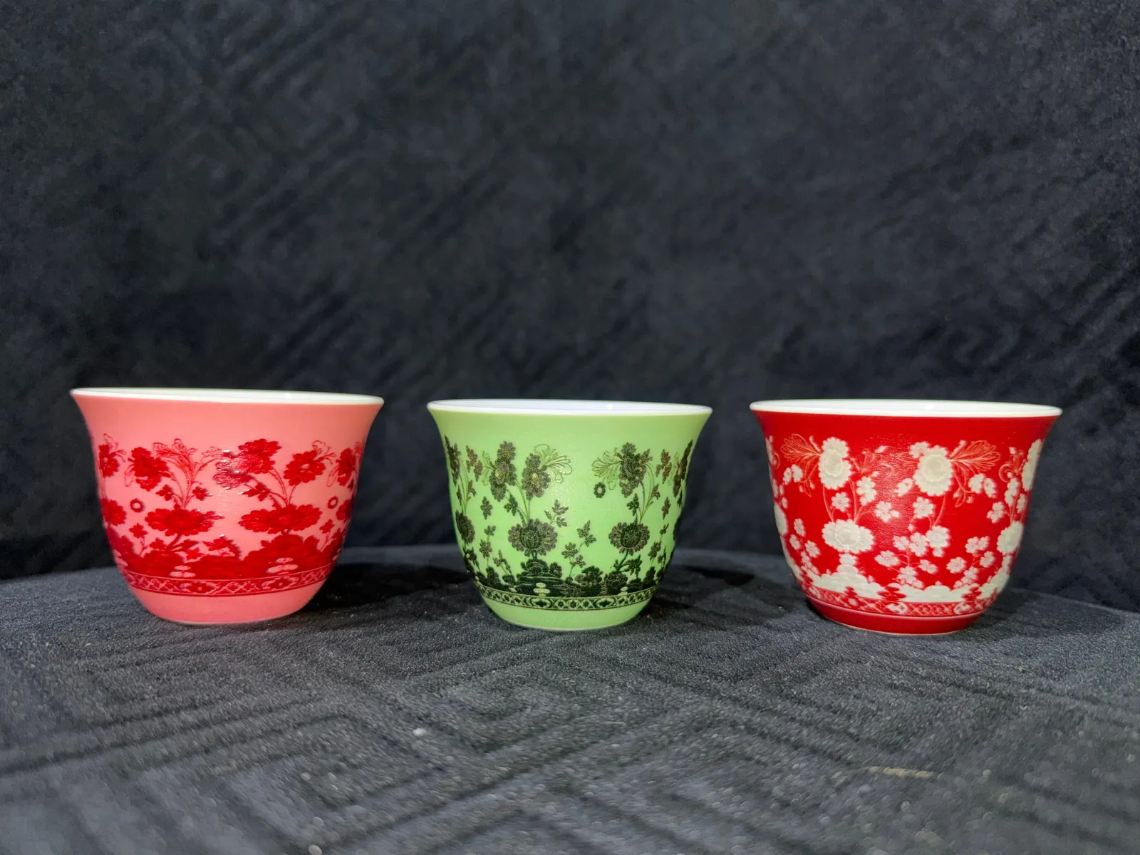 Three UV-printed porcelain qahwa cups in three colourways of one floral pattern, pink with red flowers, green with dark green flowers and red with white flowers, on a dark textured background