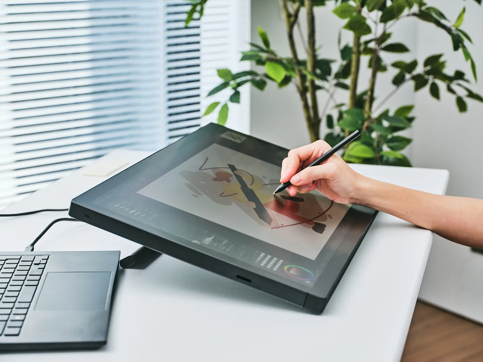 An artist drawing with the slim Wacom Pro Pen 3 on a Wacom Cintiq 24 pen display tilted on its stand beside a laptop