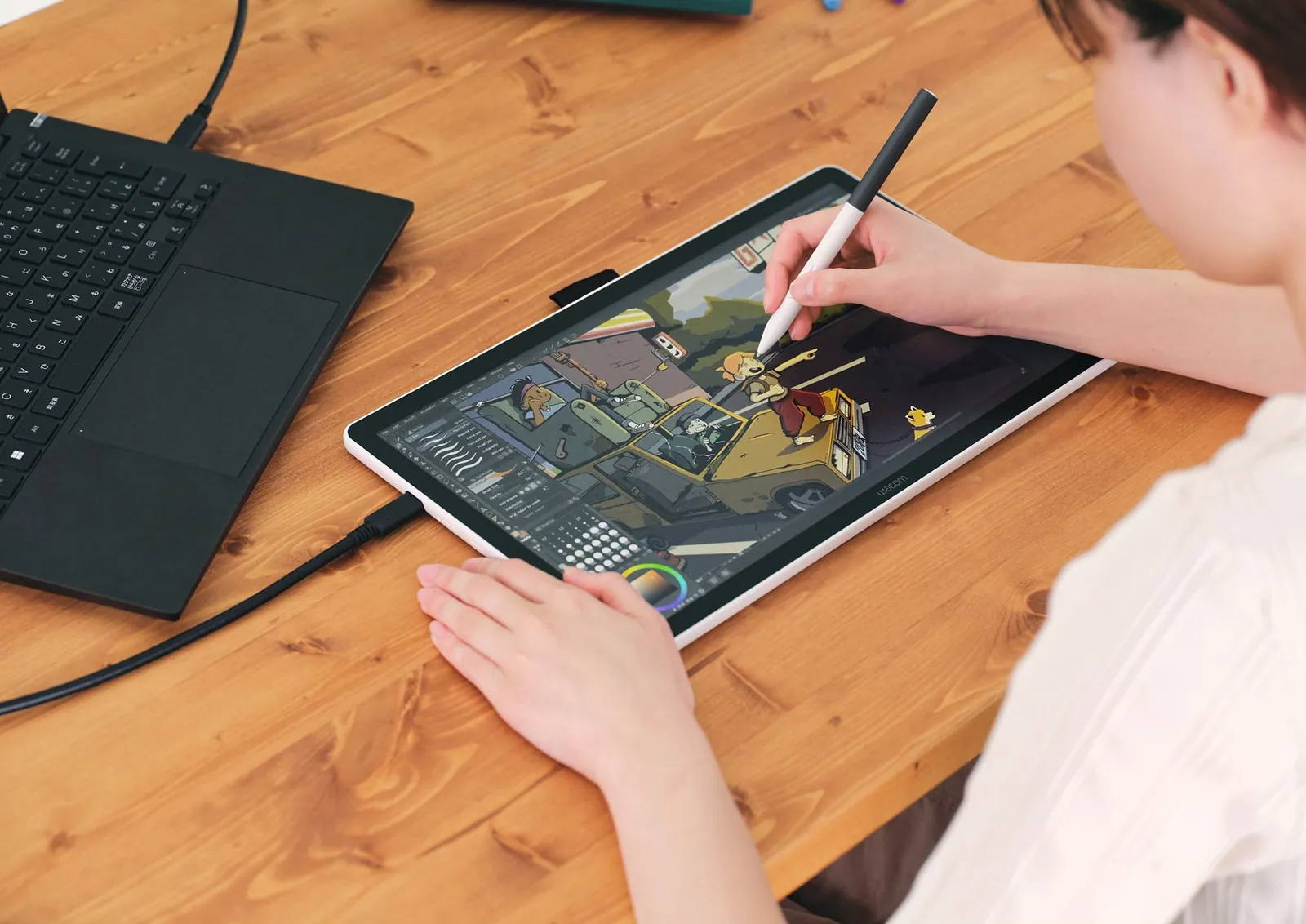 An artist drawing a comic scene with a Wacom pen on a Wacom One 14 pen display beside a laptop