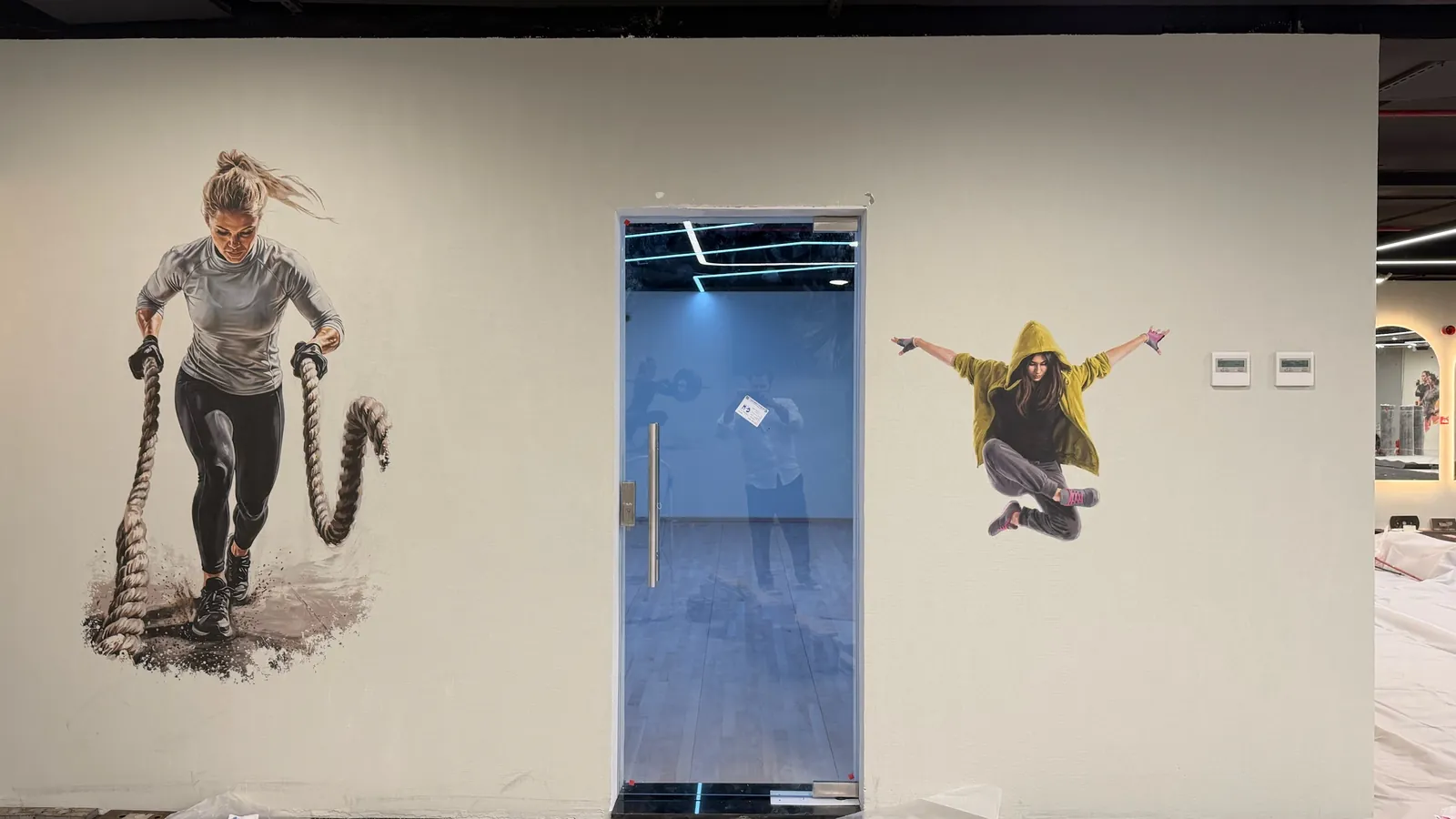 Printed gym wall showing a woman training with battle ropes on the left and a figure jumping on the right, with a glass door between them and protective sheeting on the floor